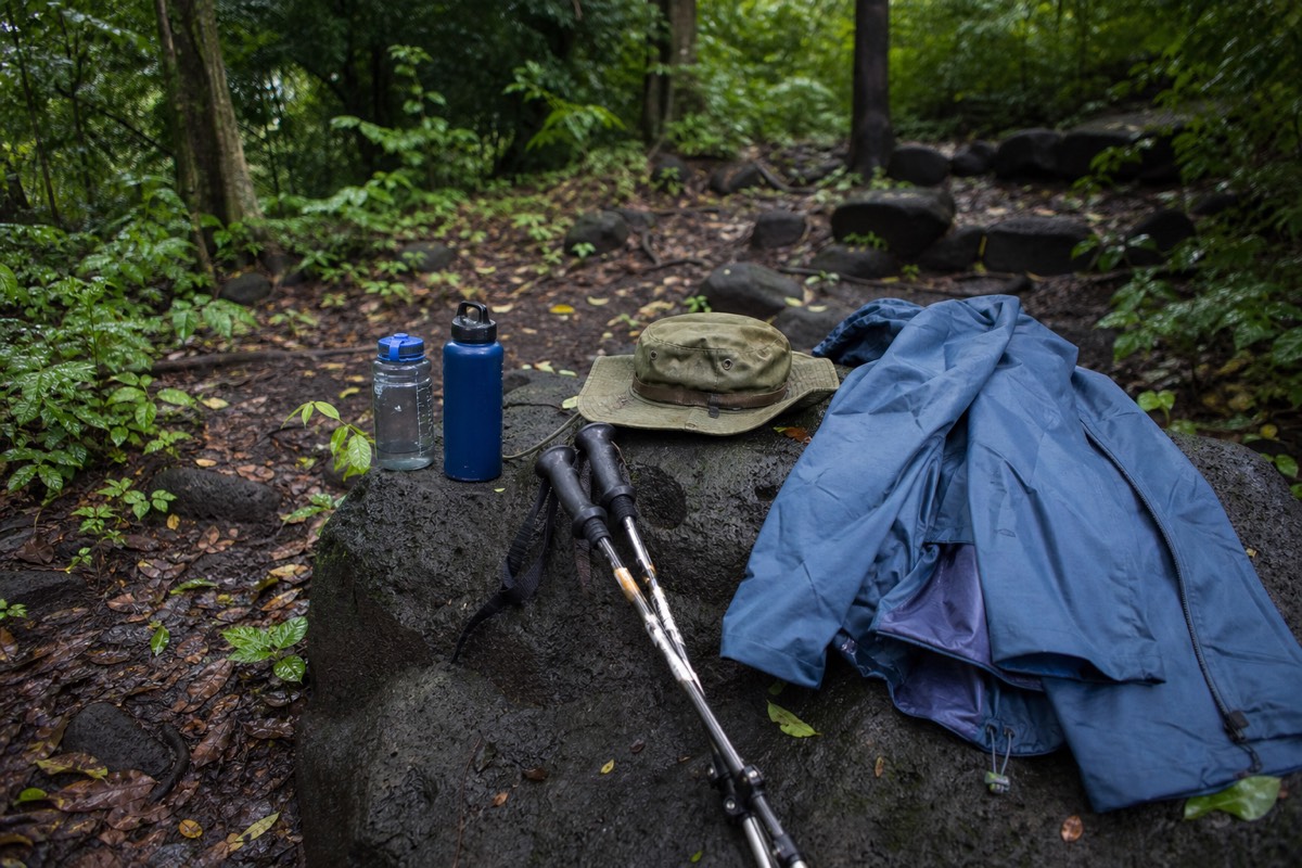 Rain shell, sun hat, water bottles and trekking poles laid out on volcanic rock at the trailhead before a green-season climb