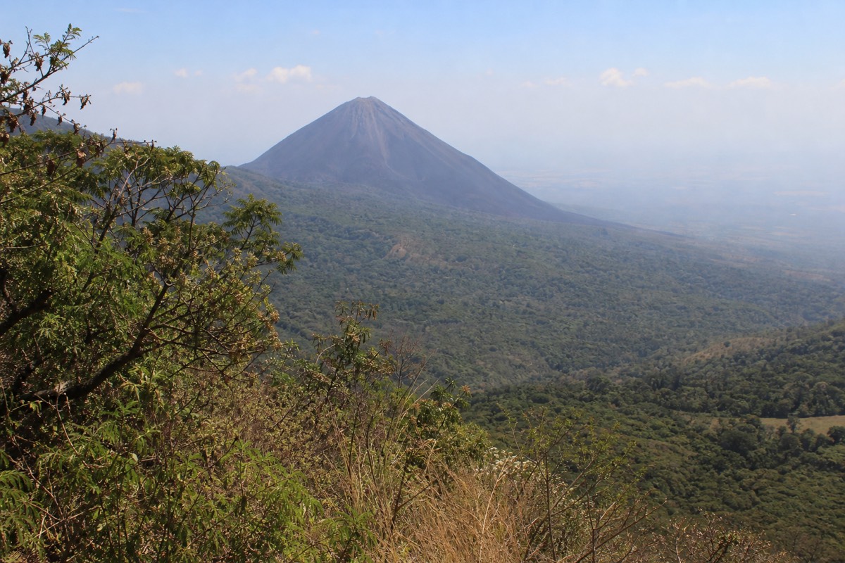 Black cinder cone of Izalco volcano seen across a green valley from the Santa Ana trail, dry-season haze on the horizon