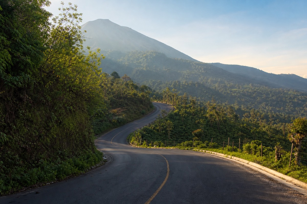 Winding paved mountain road climbing through coffee slopes and forest toward Cerro Verde National Park, volcano flank in morning haze