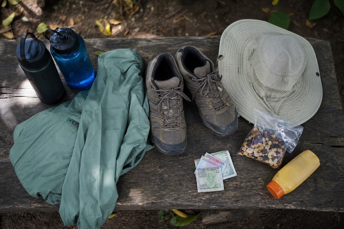 Flat lay of a Cerro Verde day kit on a wooden bench: two water bottles, light jacket, hiking shoes, hat, sunscreen, snacks and small US dollar bills