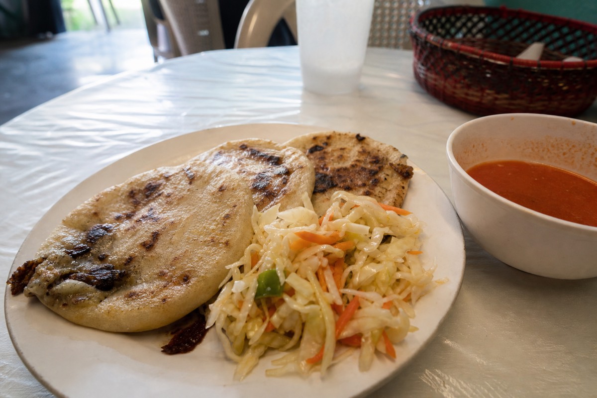 Plate of freshly griddled pupusas with curtido cabbage slaw and red salsa on a plain table at a small comedor in Santa Ana city