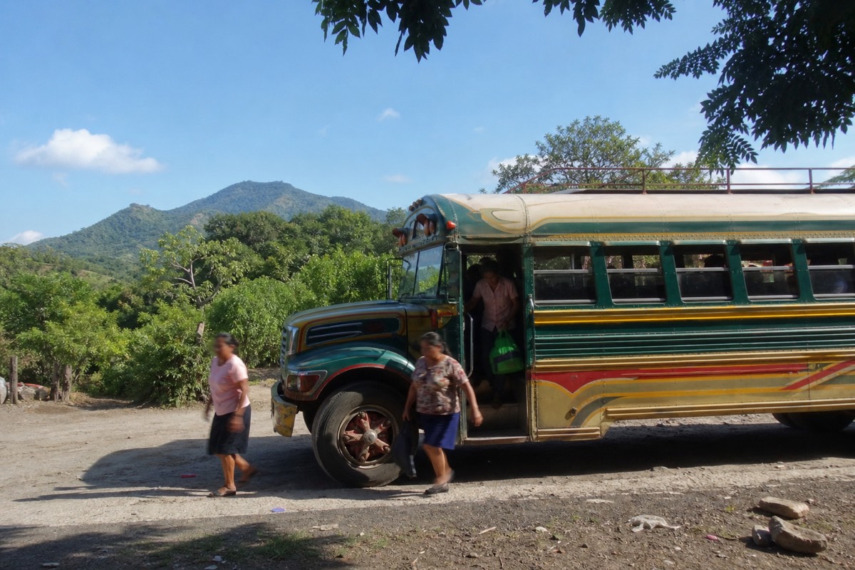 Brightly painted public bus pulling up at a dusty roadside stop in western El Salvador with green volcanic hills behind