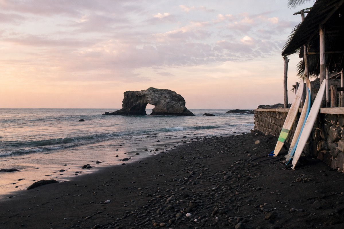 Black-sand beach at El Tunco in early morning with the rock arch offshore, gentle surf and a few surfboards leaning on a wall