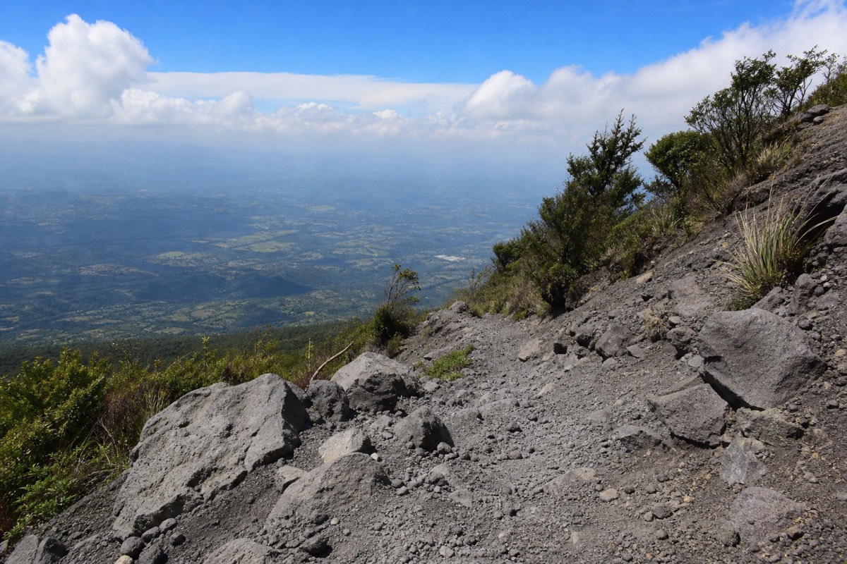Steep rocky upper trail of Santa Ana volcano under bright sun with sparse shrubs and grey volcanic gravel underfoot