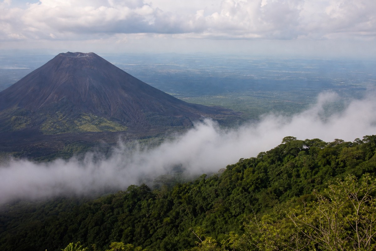 Black cone of Izalco volcano rising above dense green cloud forest seen from a Cerro Verde viewpoint under scattered cloud