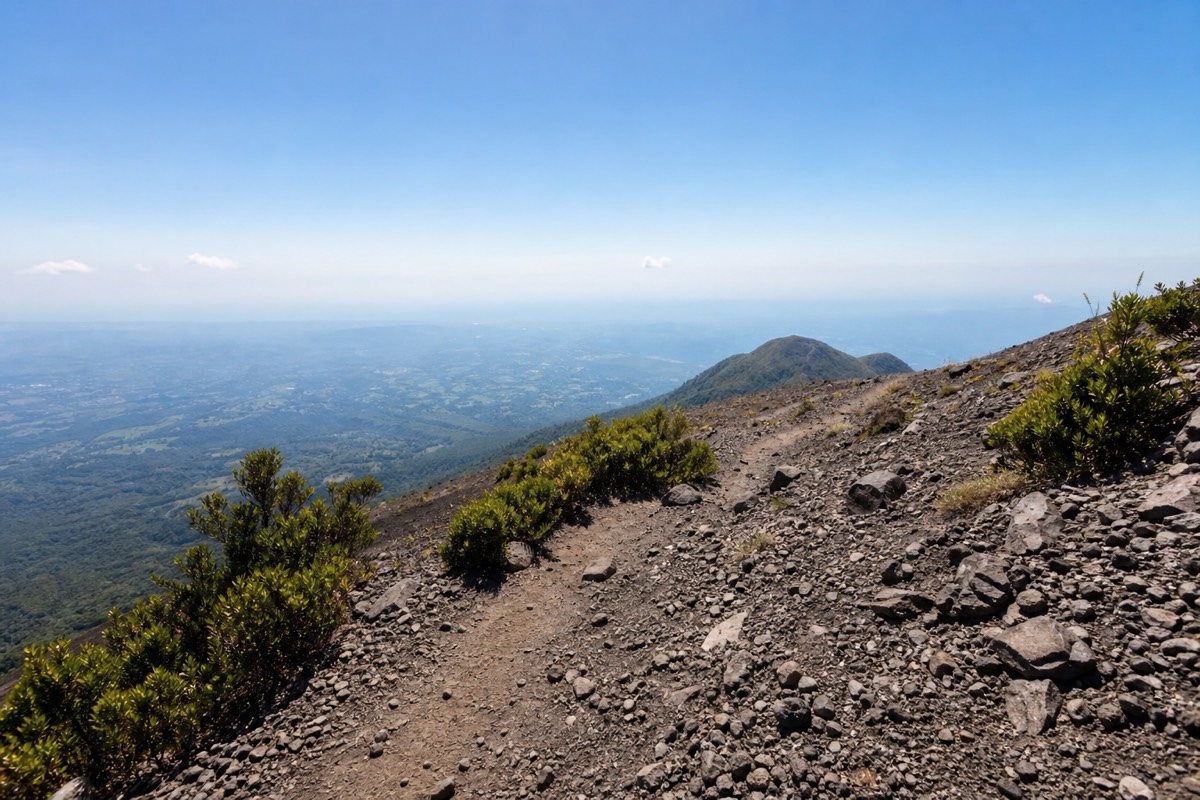 Open rocky upper section of the Santa Ana Volcano trail with loose grey stone and low shrubs, hazy lowlands far below