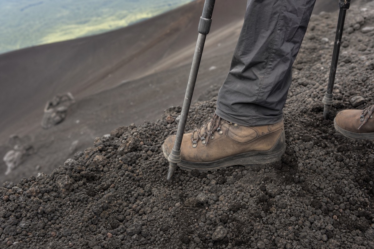 Close view of sturdy hiking boots and trekking pole tips planted on dark loose volcanic rock, steep slope falling away behind