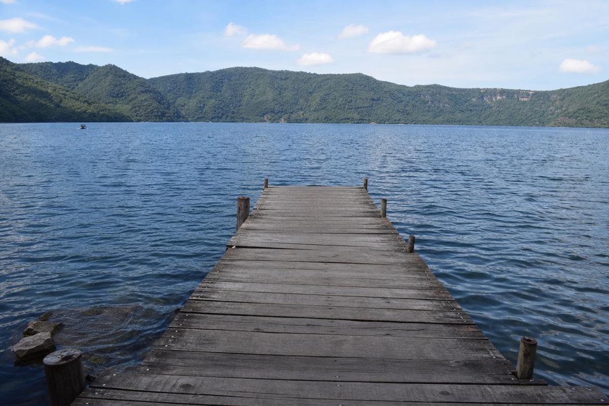 Wooden dock stretching into the calm blue water of Lake Coatepeque with green caldera walls rising on the far shore