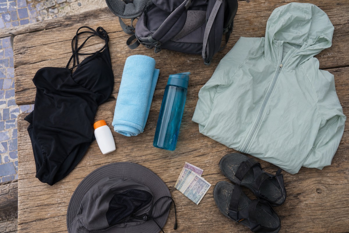 Flat lay of a daypack for the volcano and lake day: swimsuit, quick-dry towel, sunscreen, water bottle, windbreaker, sandals and small dollar bills on weathered wood