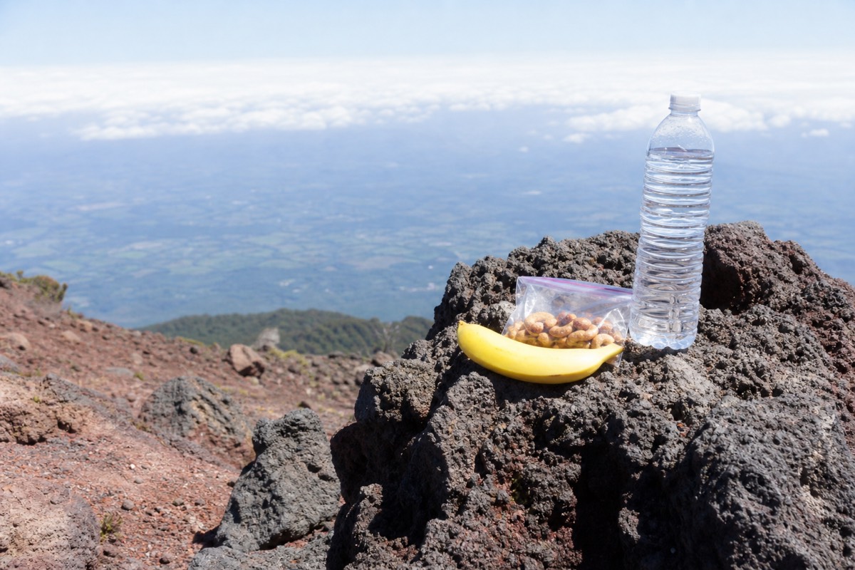 Water bottle, a banana and a small bag of nuts resting on grey volcanic rock on the upper trail of Santa Ana Volcano with hazy lowlands behind