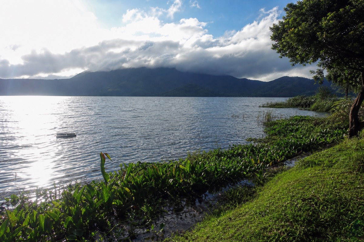 Lake Coatepeque under bright afternoon sun with wet green shoreline in the foreground and rain clouds still hanging over the volcano behind