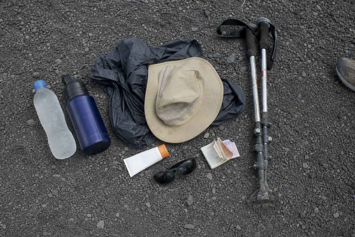 Flat lay of hiking gear for the Santa Ana summit: water bottles, windbreaker, hat, sunscreen, small US dollar bills and rented trekking poles on volcanic gravel
