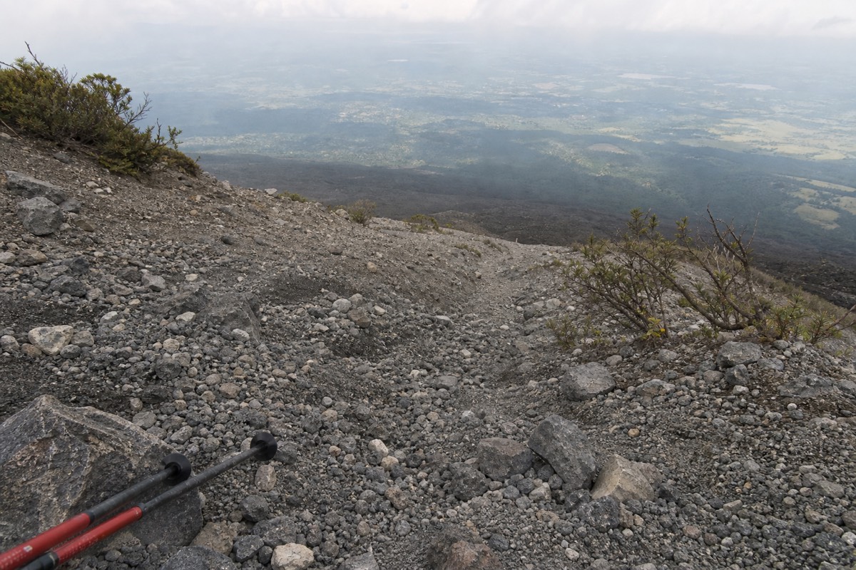 Loose grey volcanic rock and scree on the upper trail just below the crater rim of Santa Ana Volcano, trekking pole tips visible