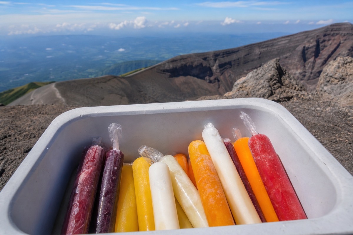 Brightly coloured fruit paletas in a vendor's cool box at the summit of Santa Ana Volcano, crater rim rock and sky behind, no readable text