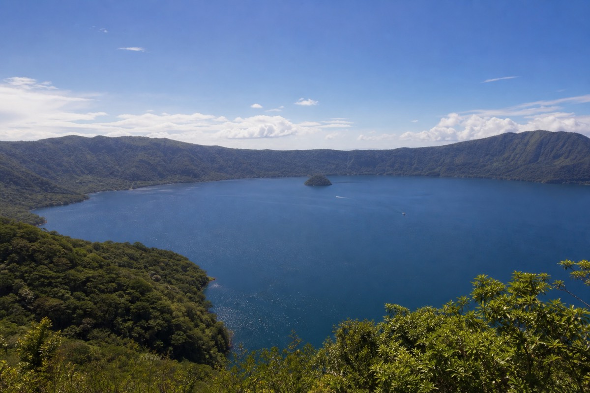 Wide view over Lake Coatepeque caldera in El Salvador, deep blue water ringed by green crater walls under midday sun