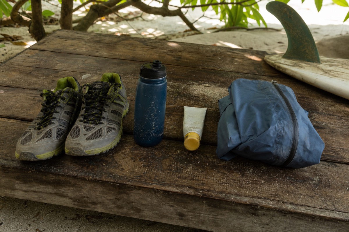 Trail shoes, a water bottle, sunscreen tube and a folded windbreaker laid out on a weathered wooden table beside a surfboard fin
