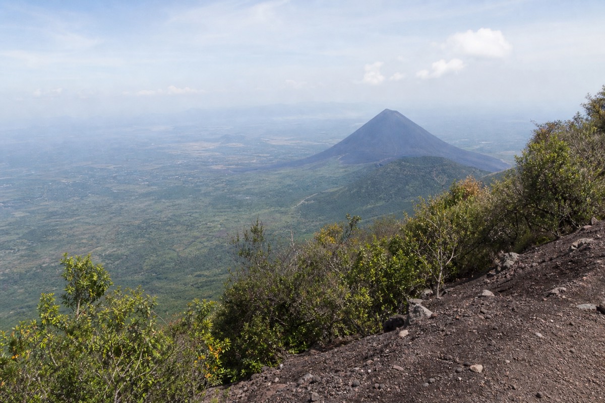 Black volcanic cone of Izalco seen across a green valley from a high trail on a neighbouring volcano under a bright hazy sky