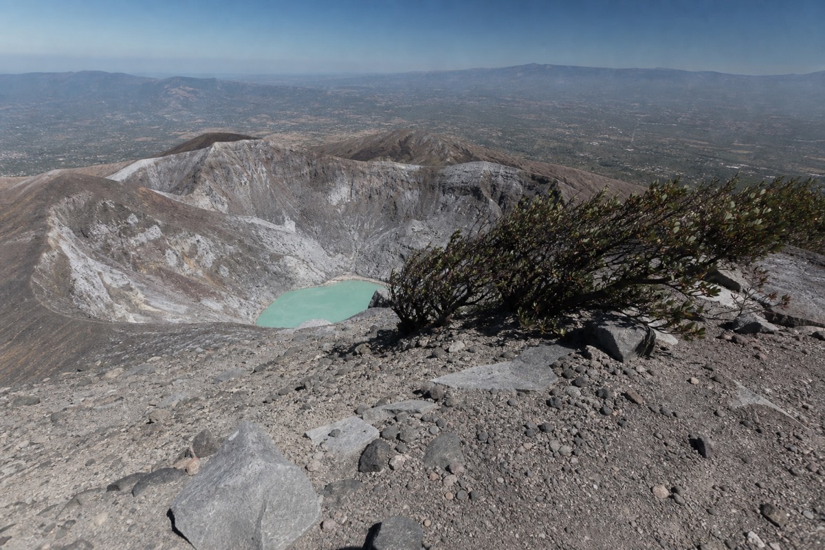 Wind-scoured rocky rim of a volcano crater in El Salvador with a pale turquoise lake far below and hardy shrubs bent by the wind
