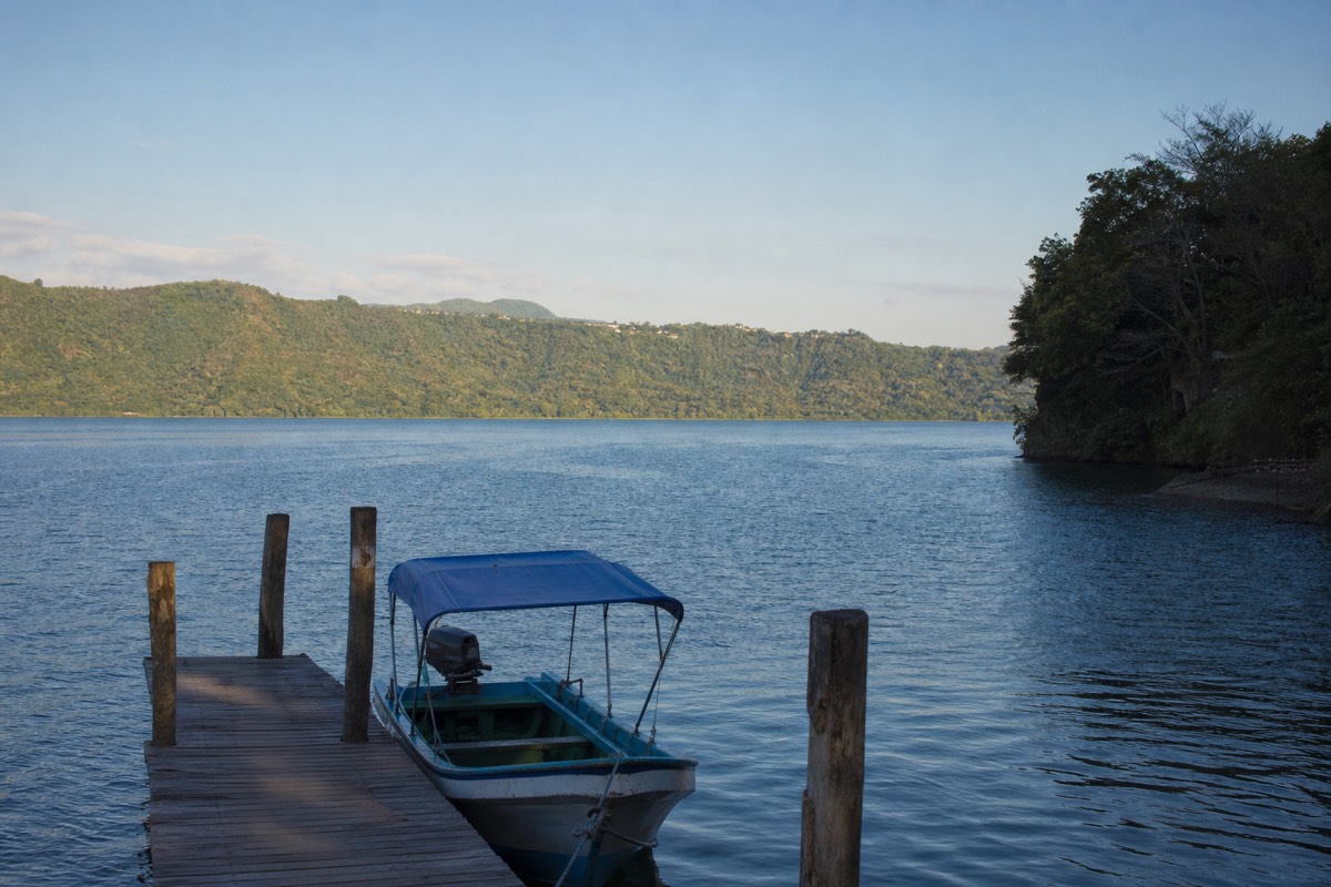 Wide blue caldera lake in El Salvador seen from a wooden dock in soft afternoon light with green hills across the water
