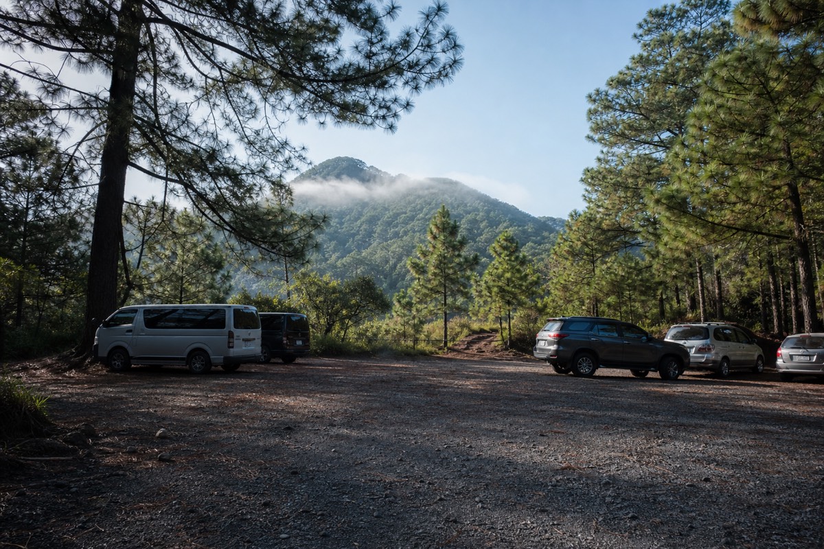 Gravel parking area at a highland volcano trailhead in El Salvador with vans and cars under pines and mist on the ridge behind
