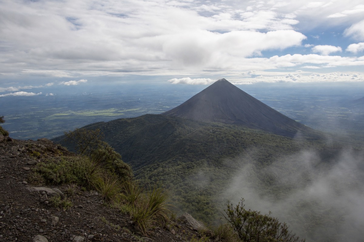 Sharp black volcanic cone rising above forested ridges in El Salvador seen from a higher trail under thin morning cloud