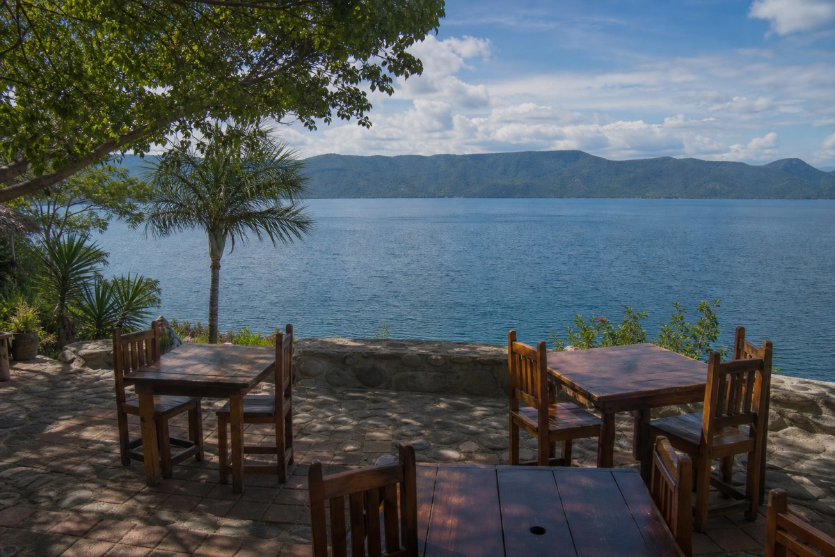Lakeside restaurant terrace over a broad blue caldera lake in El Salvador with empty wooden tables and hills across the water