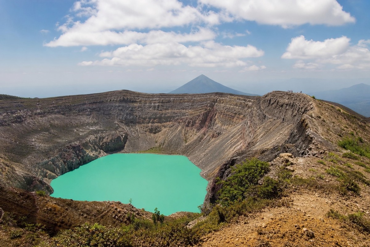Small group of hikers standing far away on the crater rim of Santa Ana volcano with the turquoise lake below and Izalco cone in the haze