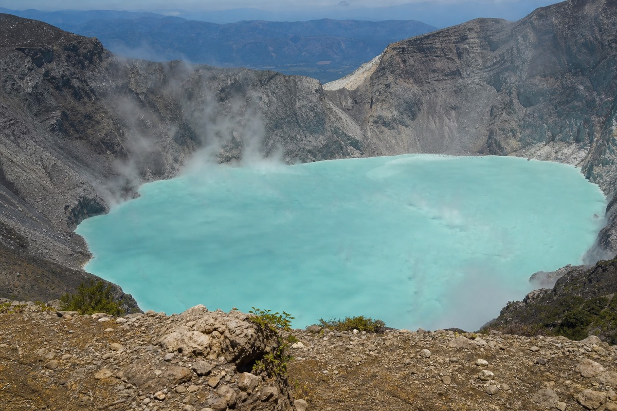 Steep grey crater walls dropping to a milky turquoise volcanic lake in El Salvador, wisps of vapour rising from the surface