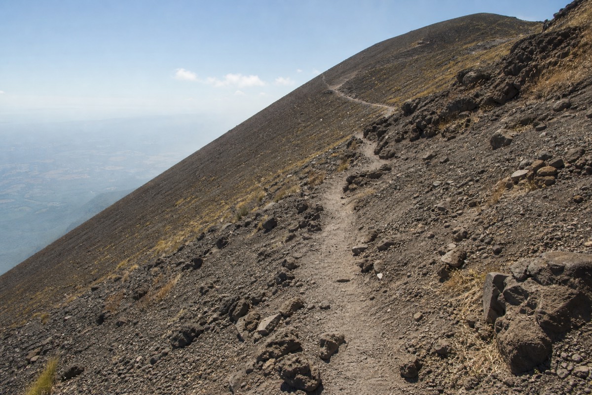 Steep loose scree and volcanic boulders on the upper trail of a big El Salvador volcano, a worn path zigzagging up toward a windy ridge