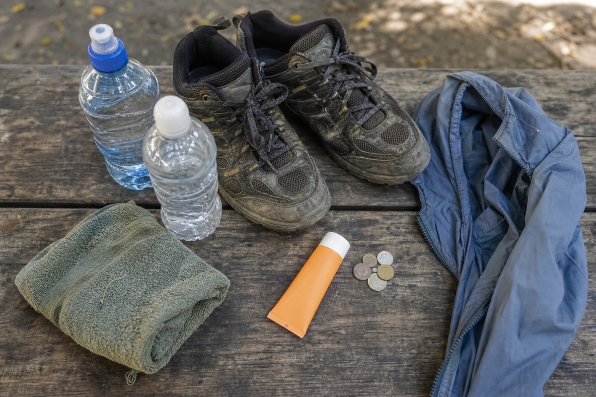 Flat lay of hiking gear for a volcano day on a wooden table: two water bottles, trail shoes, a light windbreaker, a folded towel and a small stack of coins
