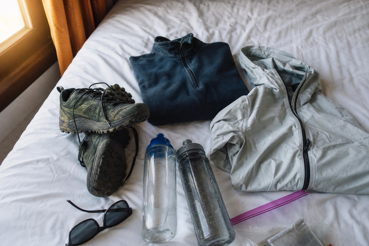 Flat lay on a hotel bed of trail shoes, a folded fleece, a light rain shell, two water bottles, sunglasses and a zip bag, packed for a one-day volcano hike