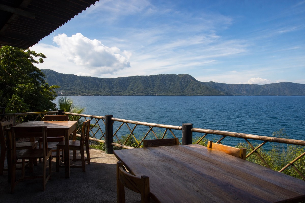Empty lakeside lunch terrace looking out over the wide blue water of Lake Coatepeque with the caldera rim green in the distance