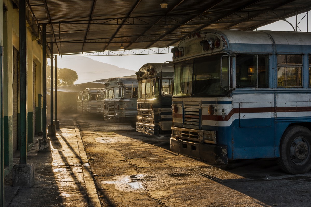 Buses lined up at an early morning terminal in Santa Ana, El Salvador, under a corrugated iron roof