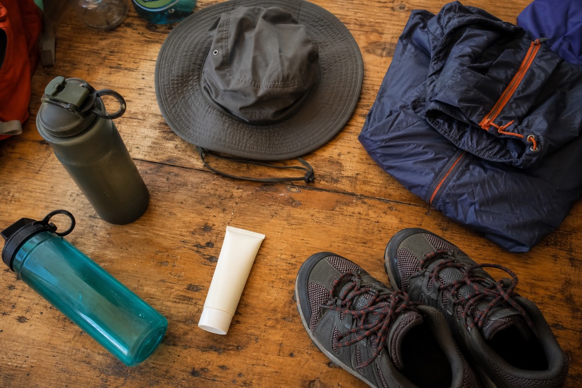 Hiking gear laid out on a wooden table: water bottles, a hat, a windproof layer, sunscreen and trail shoes