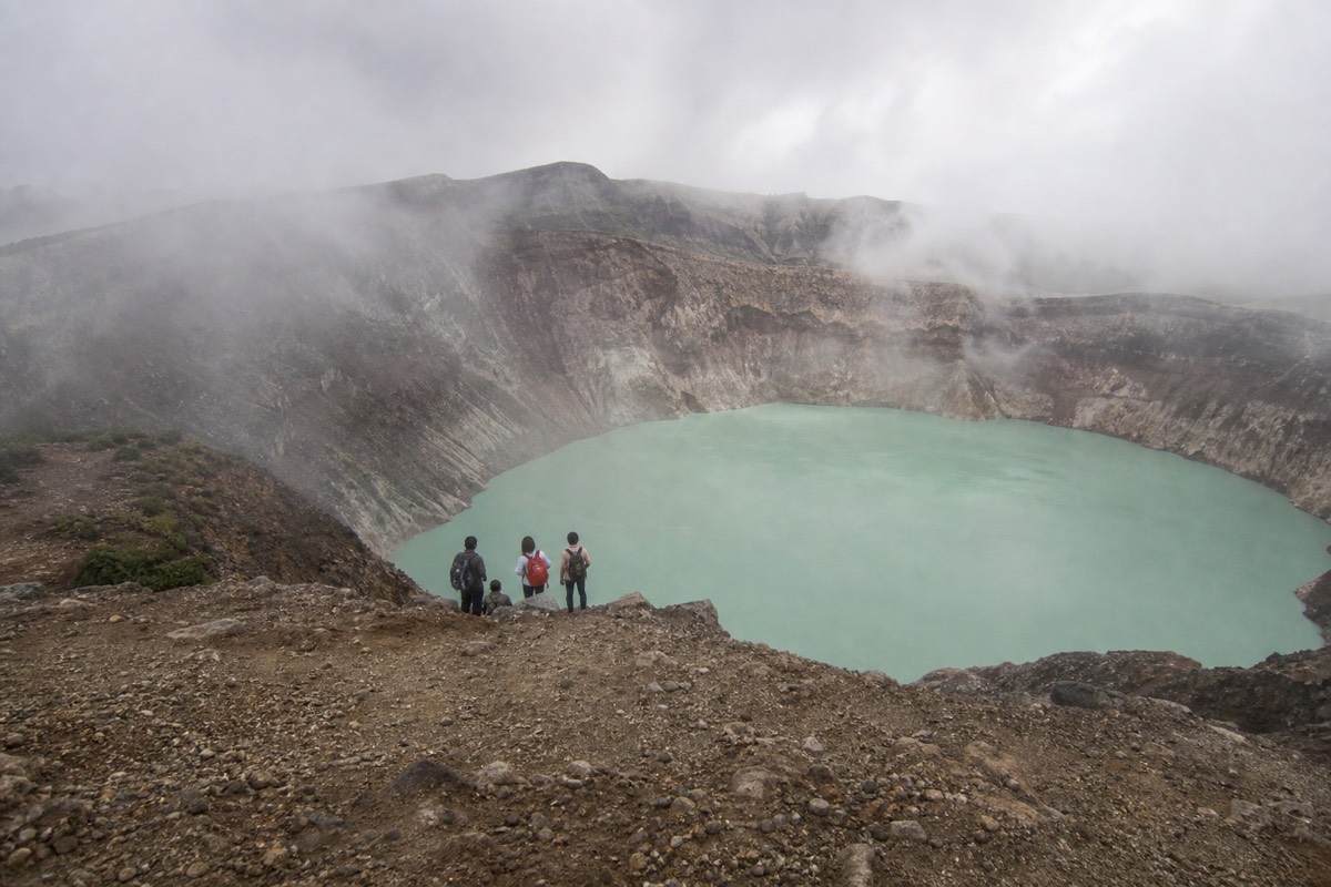 Small group of hikers seen from behind and far off, gathered together on the rocky crater rim of Santa Ana Volcano above the pale turquoise lake
