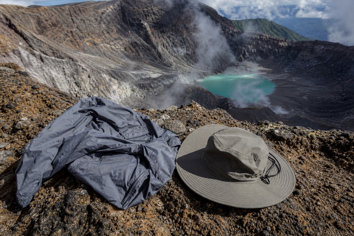A packable windbreaker and hat laid on bare rock at the windy summit rim of Santa Ana volcano with the crater lake far below