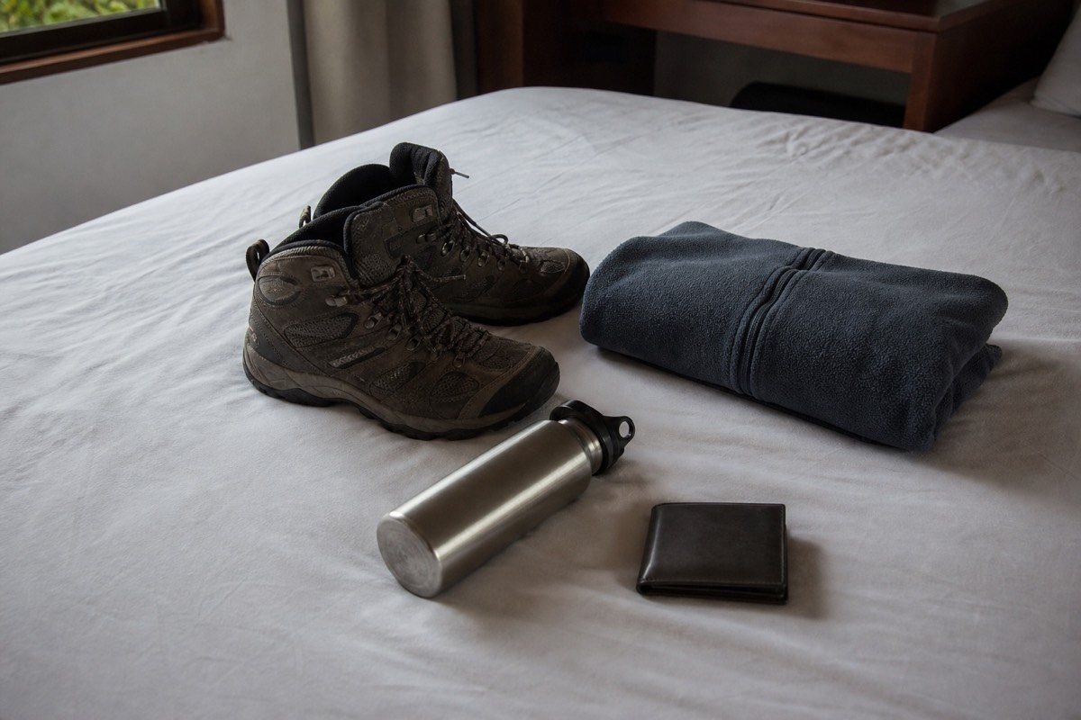 Hiking shoes, a fleece layer, a water bottle and a small wallet laid out on a plain hotel bed