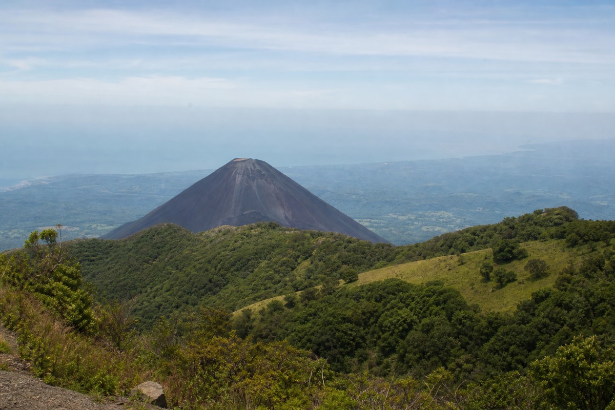 Black volcanic cone of Izalco seen across a green saddle from the upper trail of Santa Ana Volcano, sharp against hazy Pacific lowlands