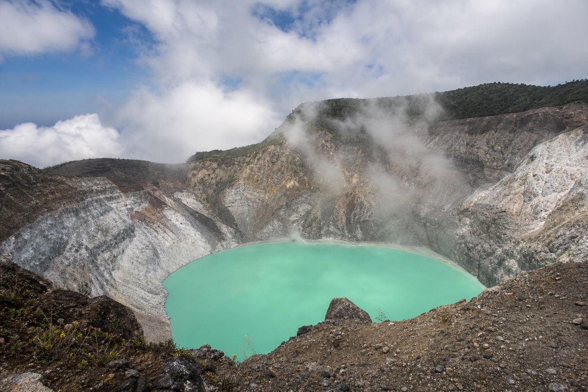 Steaming turquoise crater lake of Santa Ana Volcano seen from the rocky rim, pale sulfur-stained walls dropping to the water, thin cloud overhead